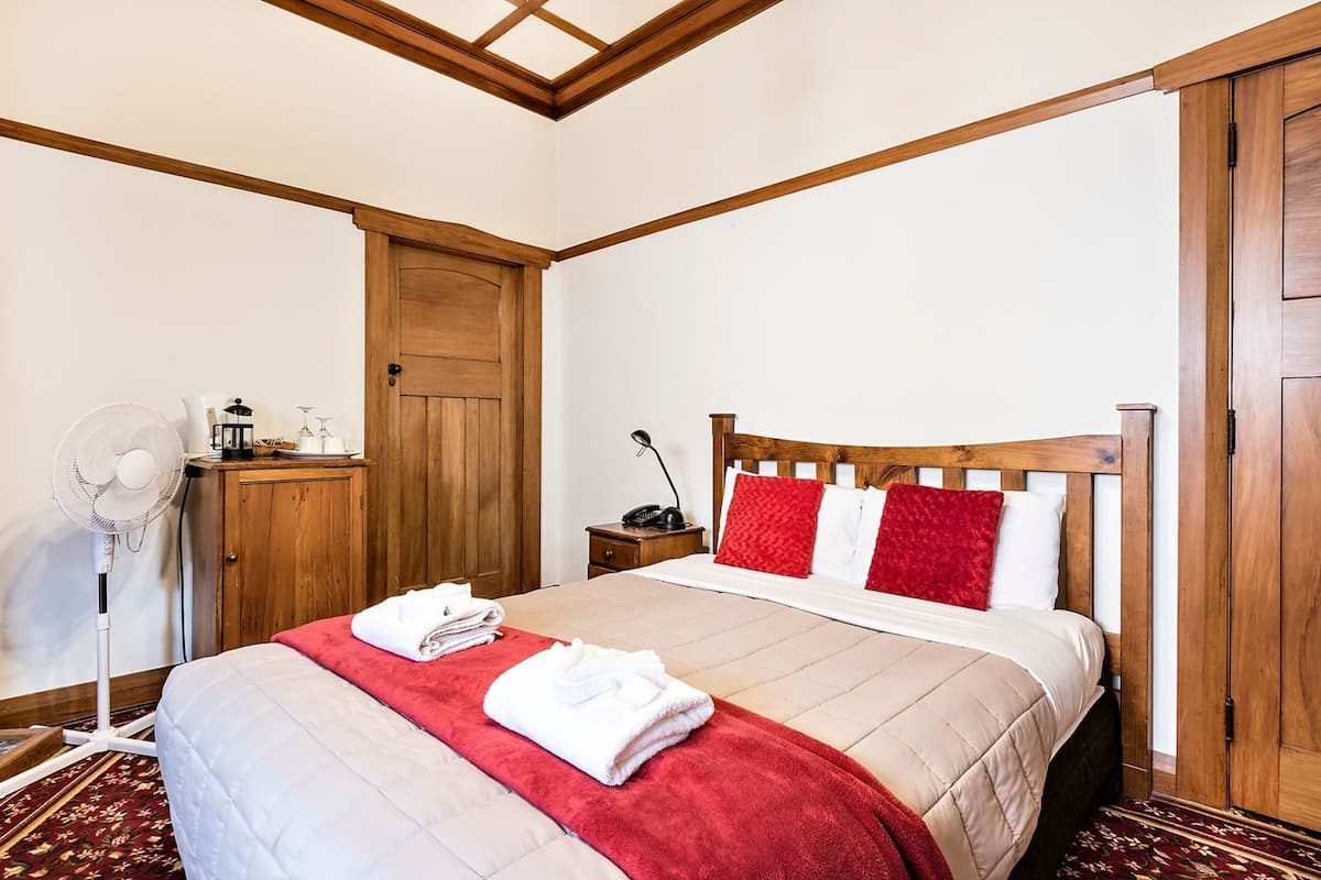 Une chambre d’hôtel avec un lit, une armoire en bois, une table de chevet, une lampe et un ventilateur sur pied.
