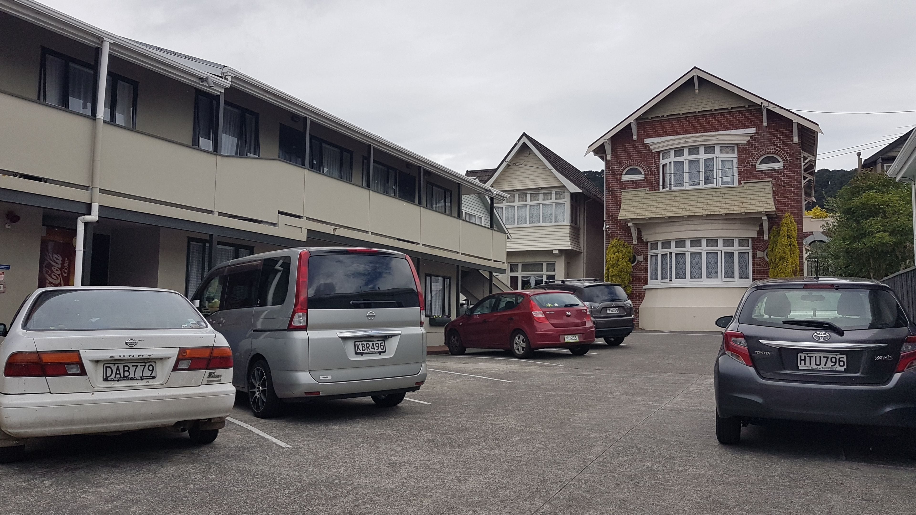 Un parking avec plusieurs voitures, un immeuble doté de nombreuses fenêtres et d’une porte de garage, et une maison en briques avec une baie vitrée.