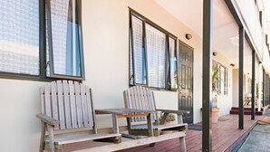 Un banco de madera con estructura metálica sobre una terraza de madera, una planta en maceta y una puerta con ventana.