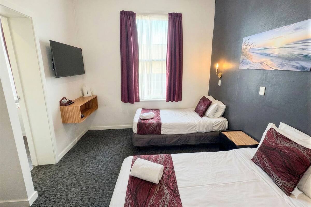 Une chambre d’hôtel avec deux lits, une télévision, une fenêtre avec des rideaux, un tableau et une table de chevet.