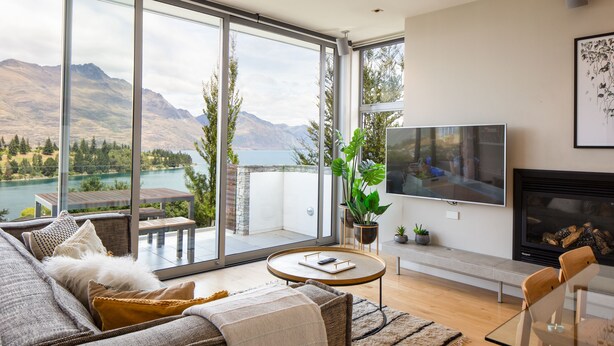 A modern living room with a large glass door leading to a balcony, a flat-screen TV, a fireplace, and a dining area. - Hello QT Pounamu (Queenstown)