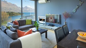 A modern living room with a large window, a fireplace, a television, and a dining area with a basket of fruit. - Hello QT Pounamu (Queenstown)