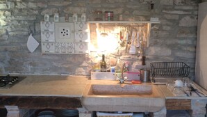 Una cocina rústica con pared de piedra, un lavamanos empotrado y estantes con utensilios de cocina y frascos.