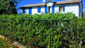 Un edificio blanco con contraventanas azules y techo de tejas, rodeado de vegetación y una cerca de metal.