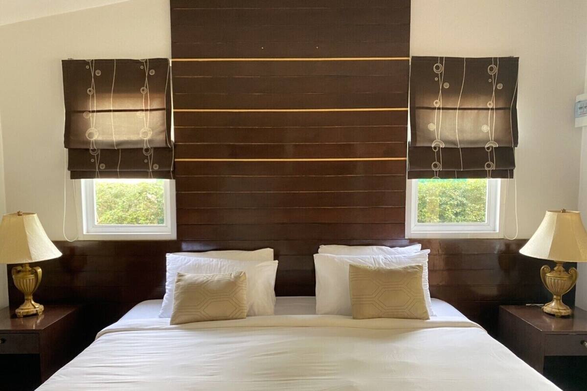 A hotel room with a large bed, two bedside tables with lamps, a dark wooden headboard, and two windows with curtains.