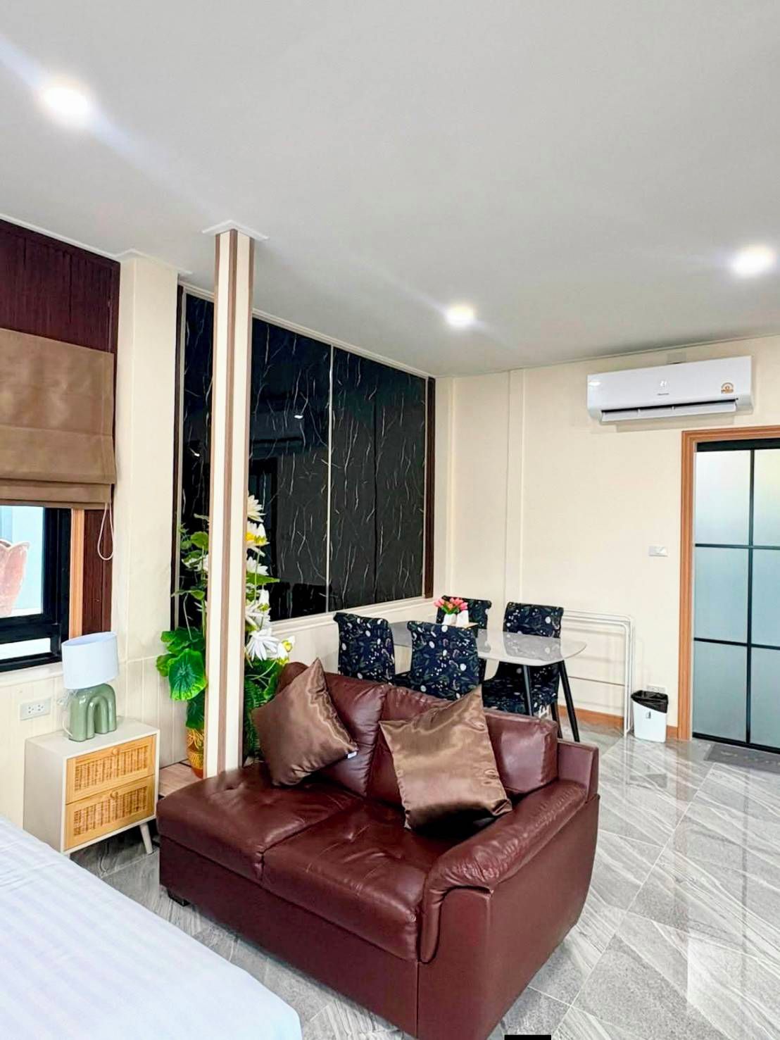 A modern living room with a brown leather sofa, a dining table with chairs, a large window with blinds, and an air conditioning unit.