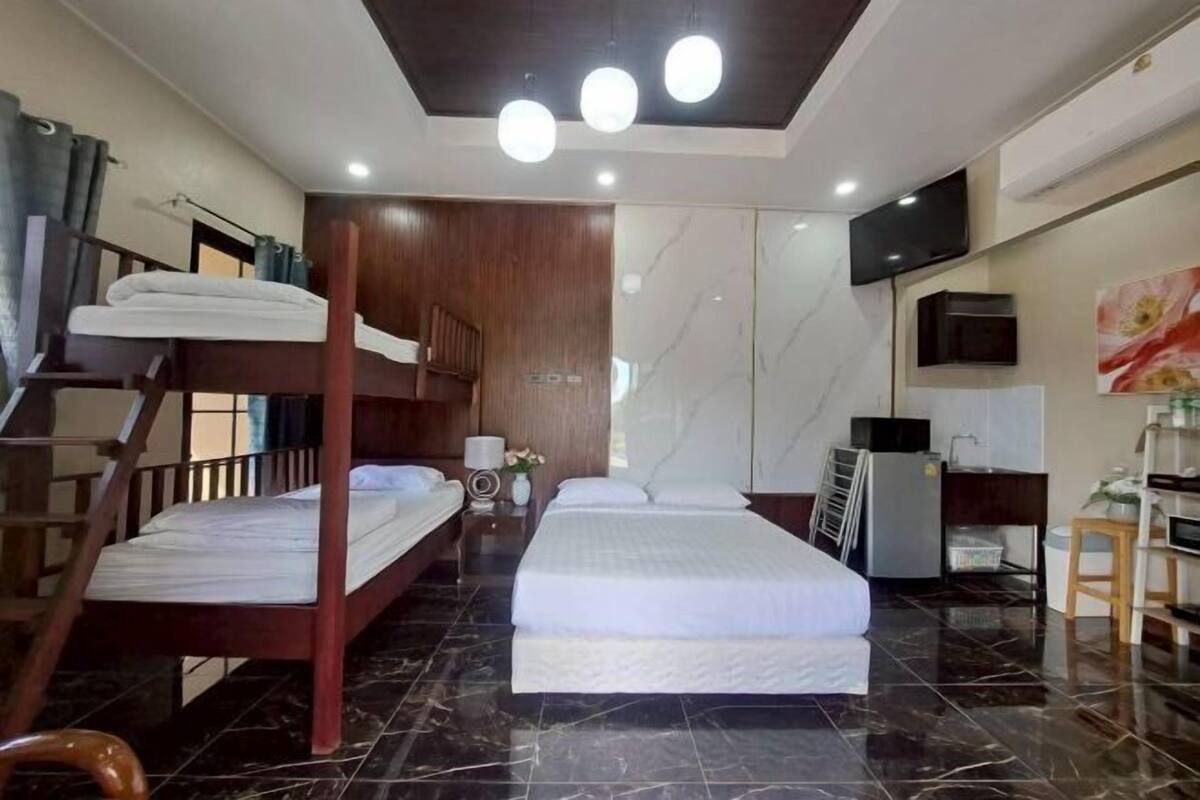 A hotel room with a bunk bed, a single bed, a flat-screen TV, a desk with a chair, and a refrigerator.