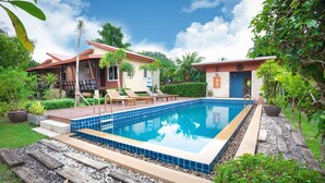 A rectangular swimming pool with blue tiles, surrounded by a wooden deck and steps leading to a covered patio area with lounge chairs. - Siray Green Resort (Ratsada)