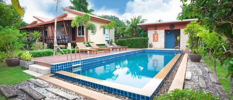 A rectangular swimming pool with blue tiles, surrounded by a wooden deck and steps leading to a covered patio area with lounge chairs.