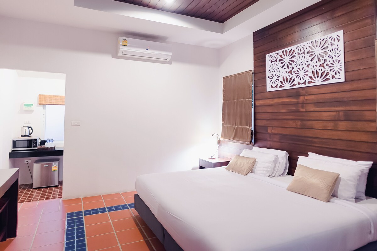 Ein Hotelzimmer mit Bett, Nachttisch, Wand-Klimaanlage, einer Dekoration und einer kleinen Küchenzeile im Hintergrund.