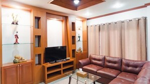 A living room with a brown leather sofa, a glass coffee table, a flat-screen TV on a wooden stand, and a ceiling fan. - Siray Green Resort (Ratsada)