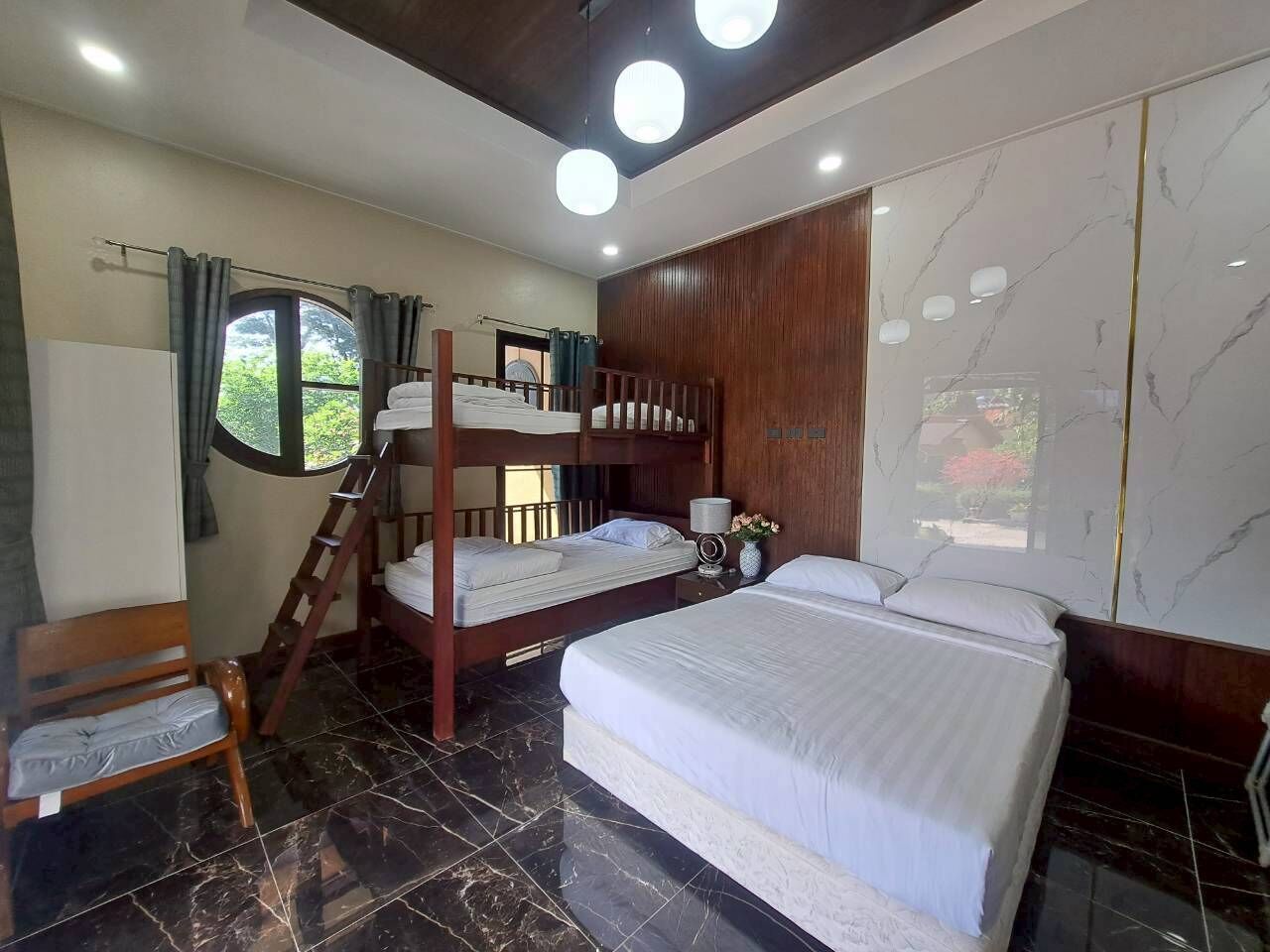Une chambre avec un lit superposé, un lit simple, une chaise en bois, une échelle, une table de chevet et une fenêtre avec des rideaux.