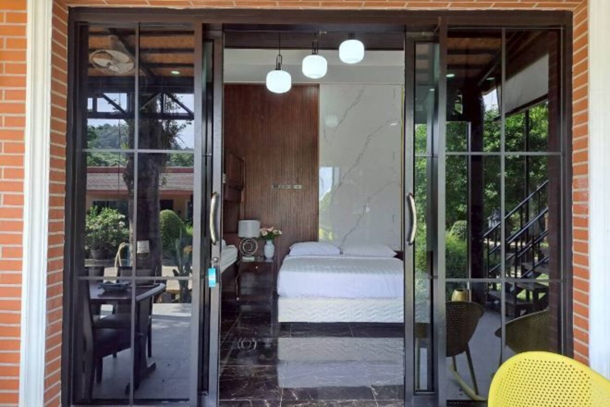 Una habitación con cama, una mesita, una silla y un vidrio que da a un espacio exterior con otra silla y una mesa.