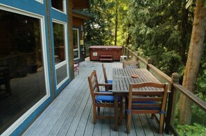 Una terraza de madera con una mesa, sillas, un jacuzzi y vistas a un bosque denso.