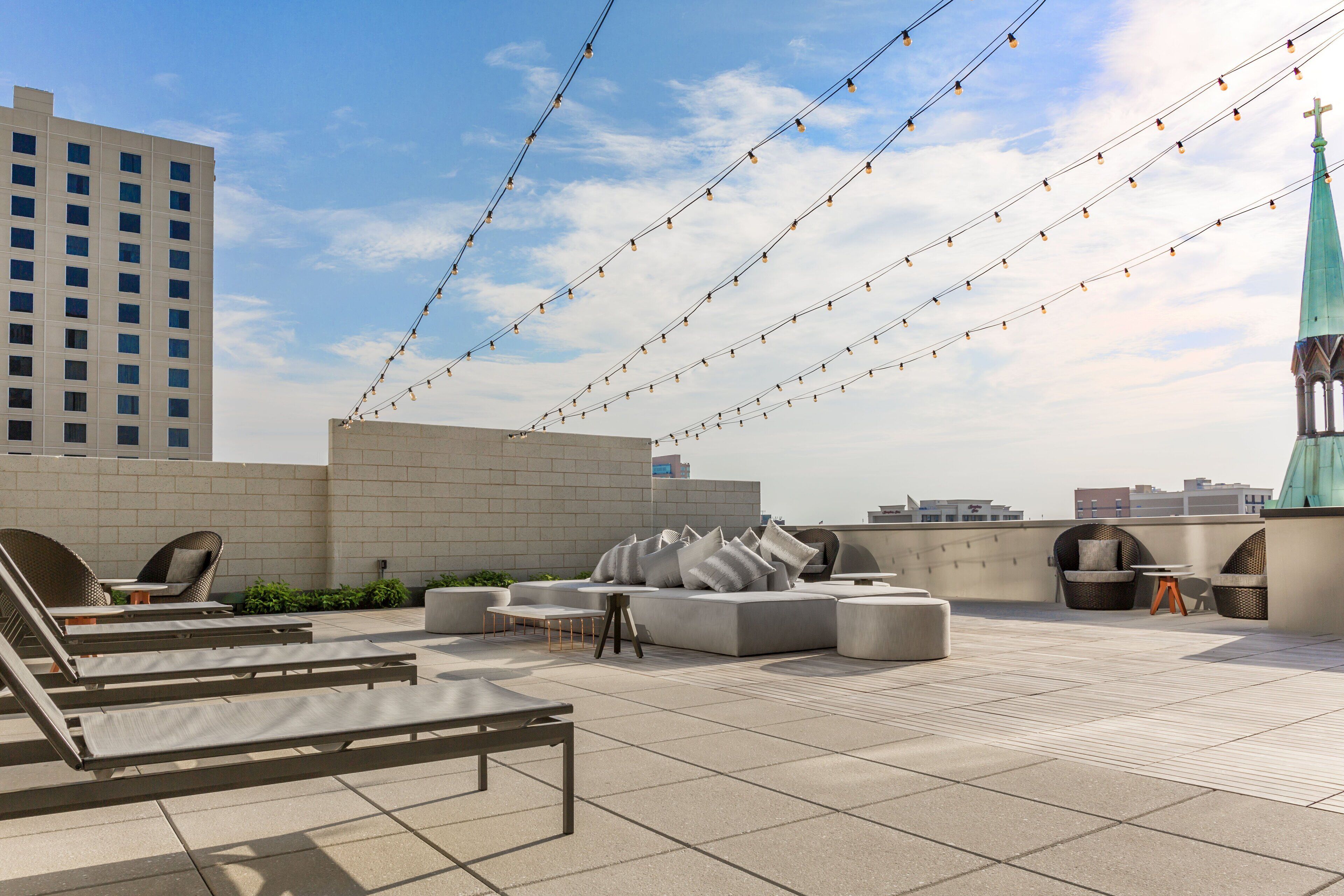 A rooftop terrace with lounge chairs, a sofa, and string lights against a cityscape backdrop.