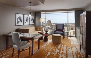 Habitación de hotel con un ventanal grande, un sofá, una mesa de comedor, una silla, una lámpara y un armario.