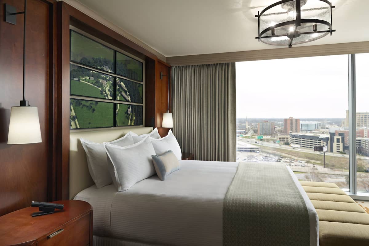 A hotel room with a large bed, a nightstand, a lamp, a view of the city through large windows, and a painting on the wall.