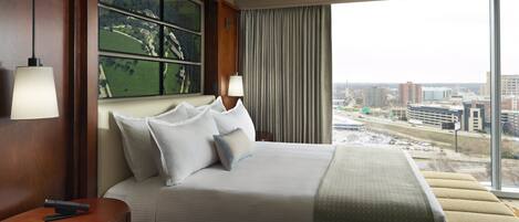 Habitación de hotel con una cama grande, una mesita de noche, una lámpara, vistas de la ciudad a través de amplios ventanales y un cuadro en la pared.