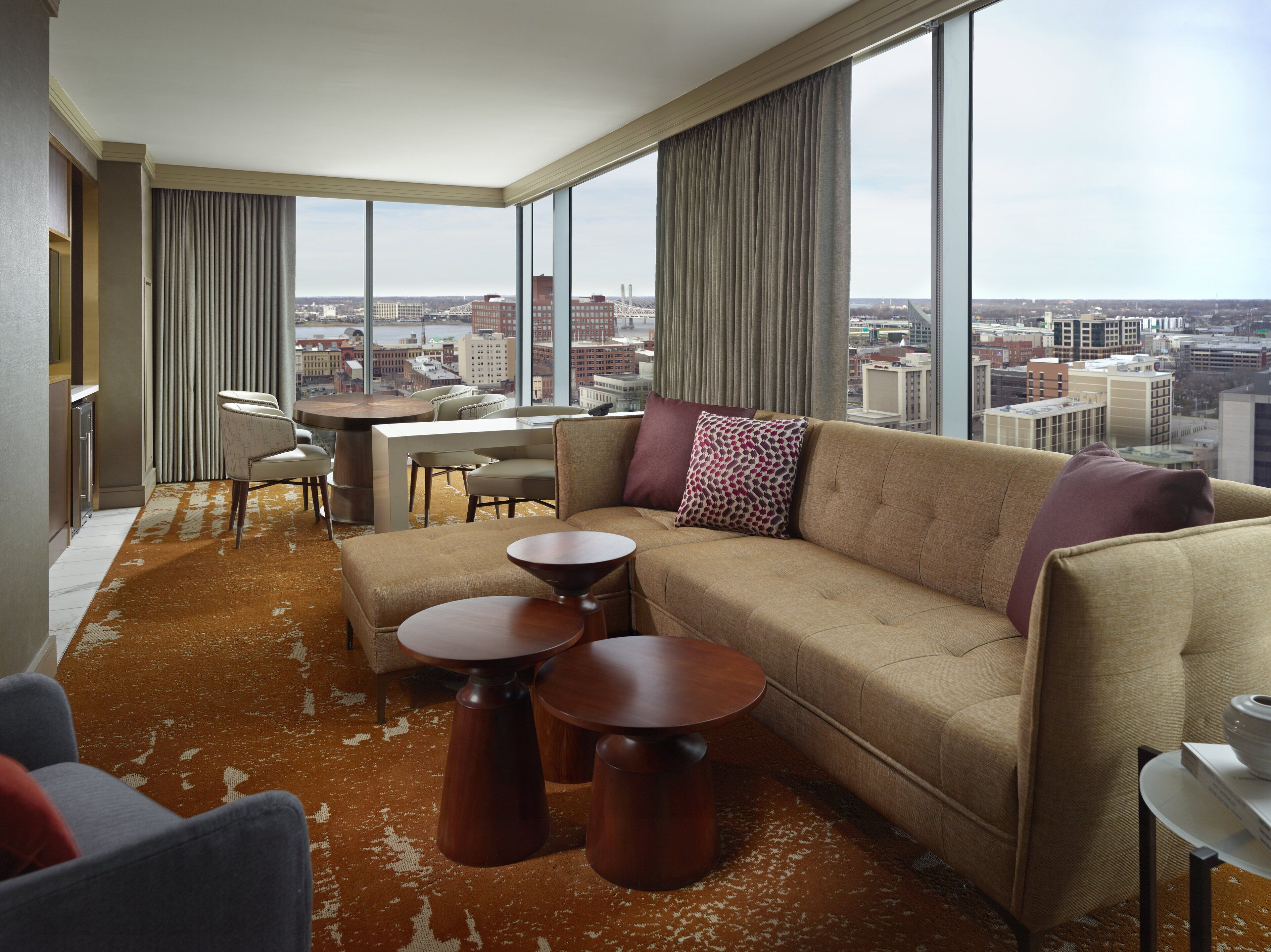 A modern hotel room with a large window offering a city view, a sofa, two small tables, and a chair.