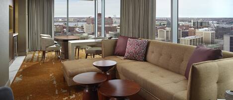 A modern hotel room with a large window offering a city view, a sofa, two small tables, and a chair.