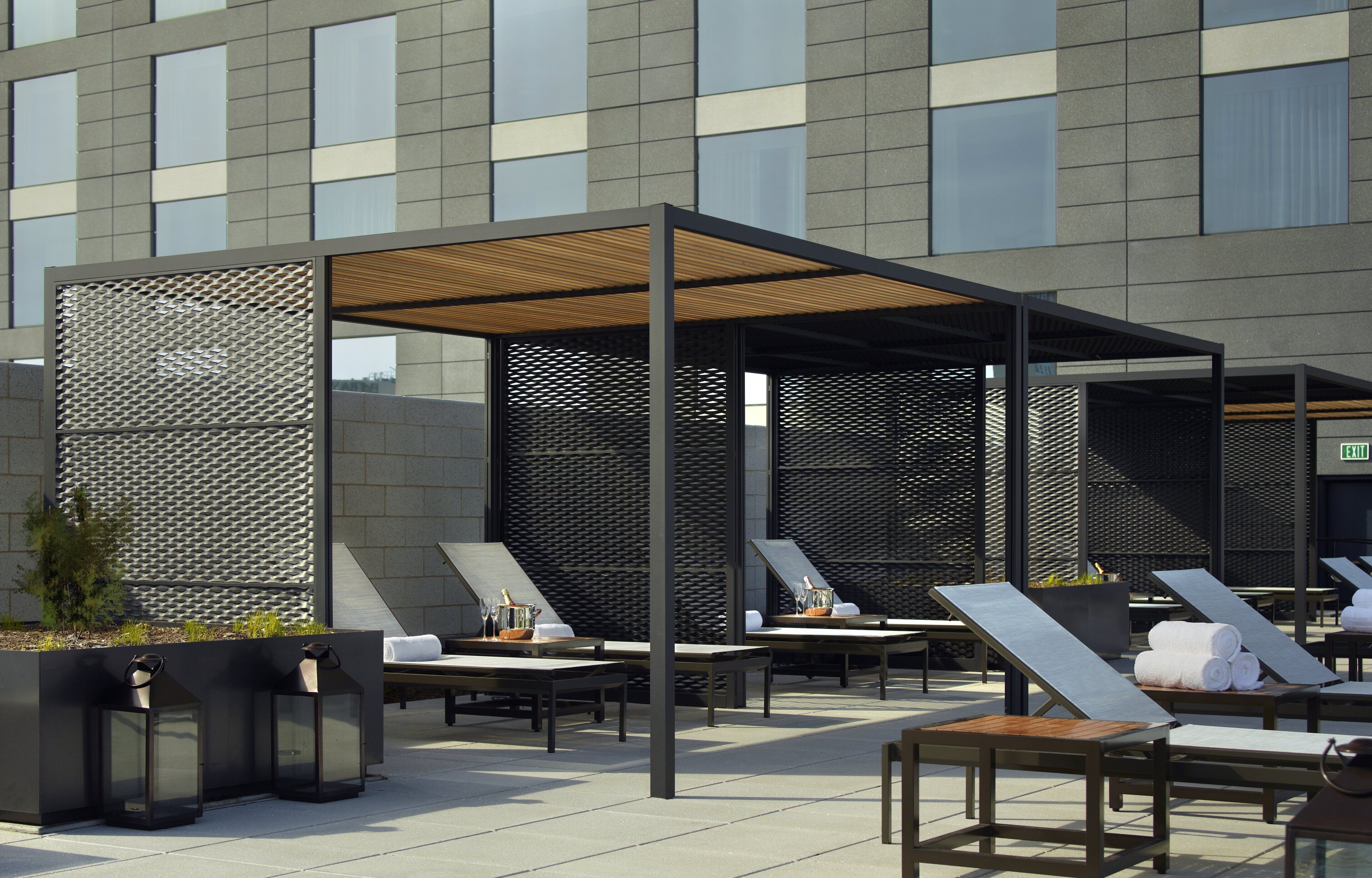 Un área de estar al aire libre con sillones de sol y mesas, una pérgola de madera y un edificio moderno al fondo.