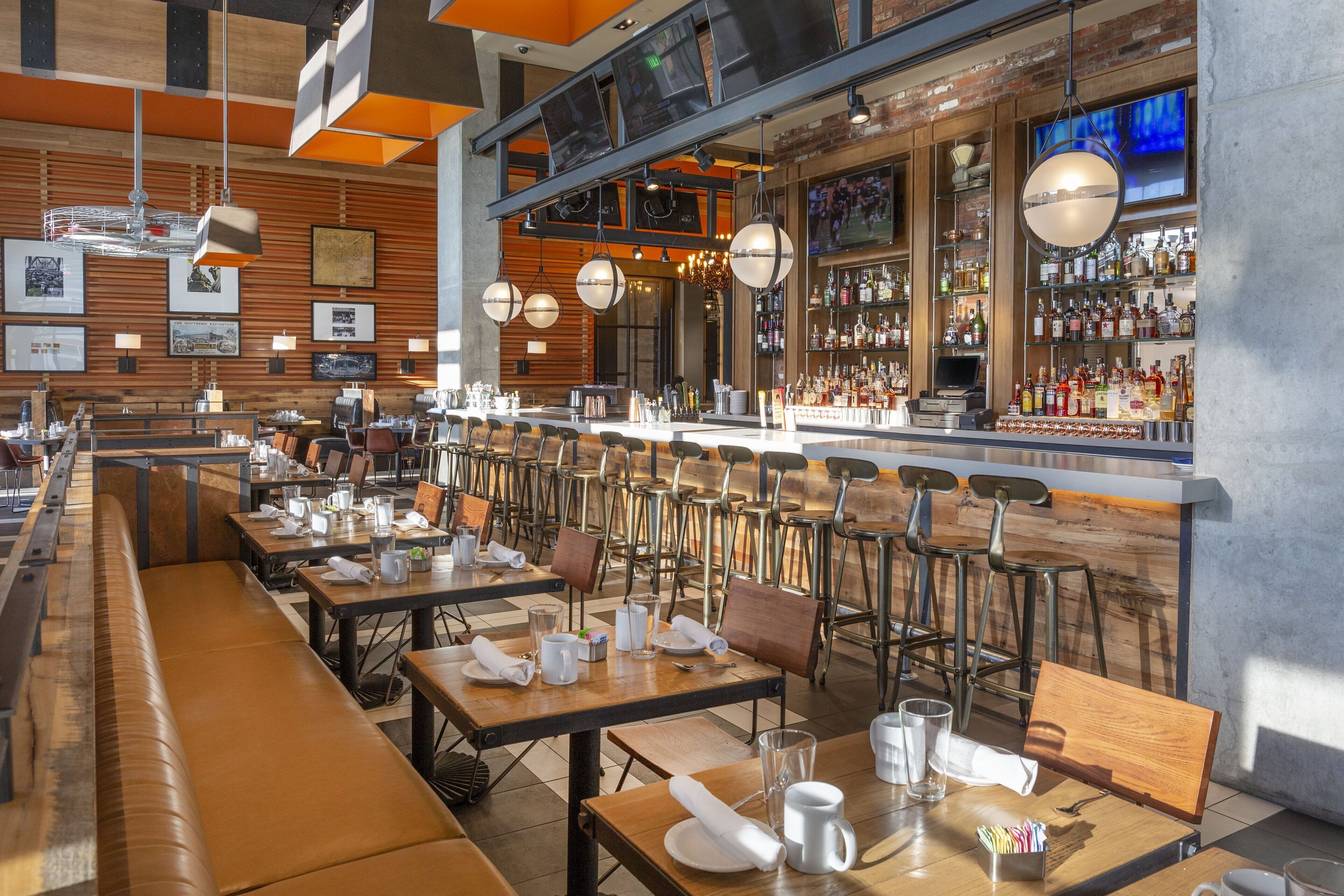 Un restaurante moderno con varias mesas y cabinas, una zona de bar con estantes llenos de botellas y lámparas colgantes.