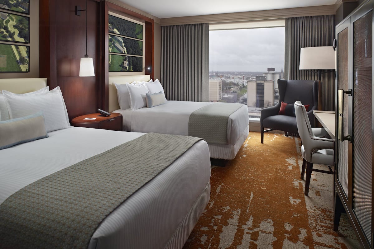 A hotel room with two beds, a desk, a chair, a lamp, and a large window with a city view.