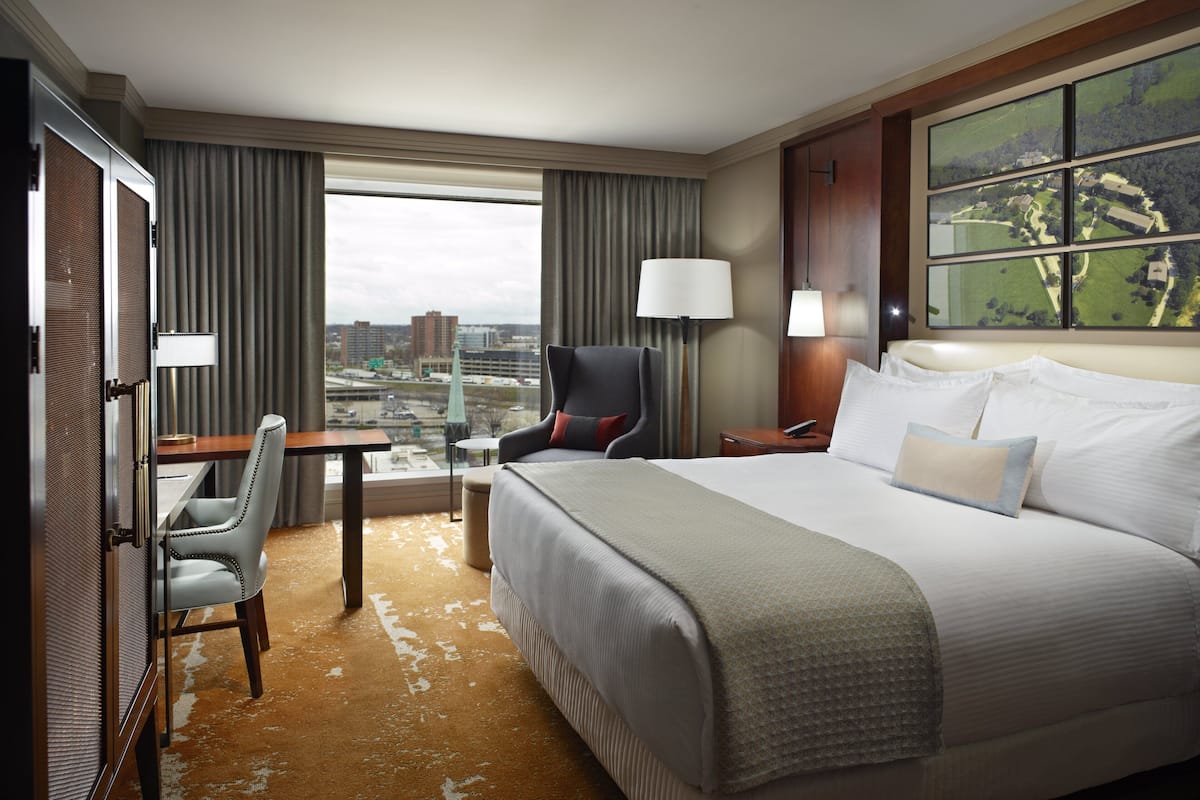 A hotel room with a large bed, a desk, a chair, and a view of a cityscape through the window.