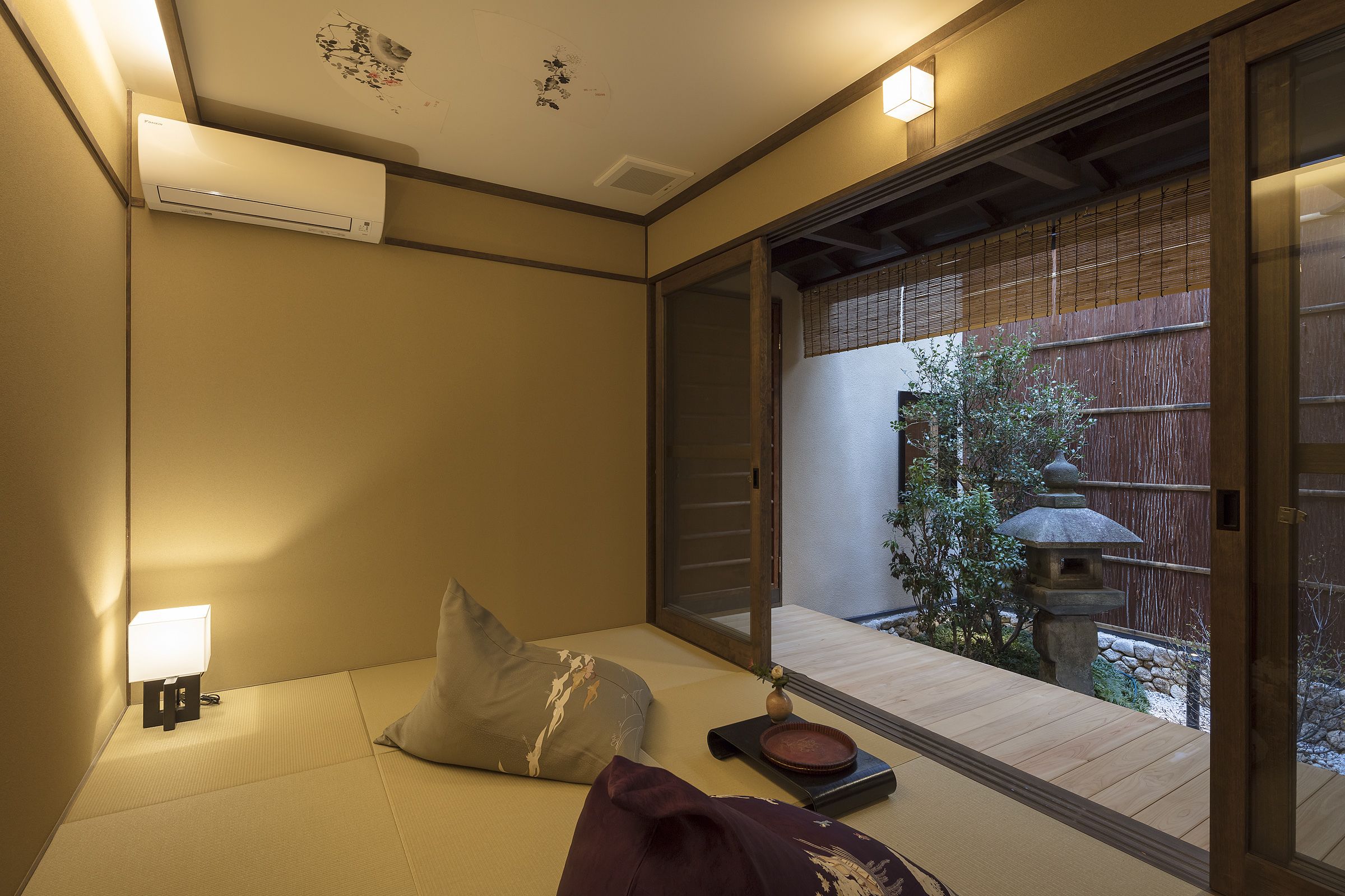 A traditional Japanese-style room with tatami flooring, a low table with a lantern, and a sliding door leading to an outdoor area with a wooden fence and a stone lantern.