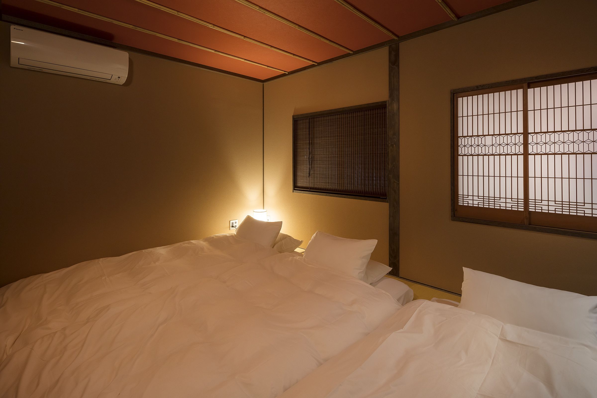 A hotel room with a bed, a wall-mounted air conditioner, a window with a grid pattern, and a bedside lamp.