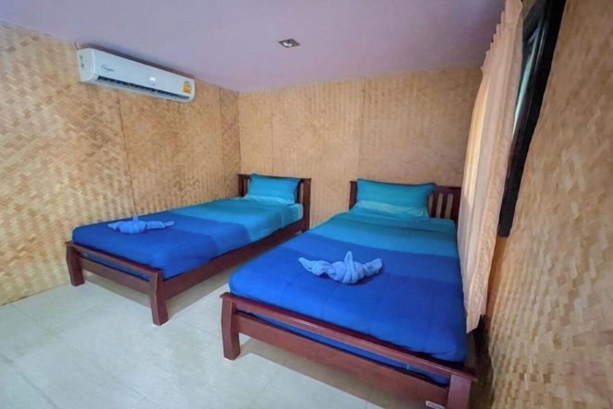 Two single beds with wooden frames and blue bedding, an air conditioning unit on the wall, and a window with curtains.