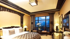 Habitación de hotel con una cama grande, un escritorio con silla, televisor y vistas a la ciudad a través de amplios ventanales.