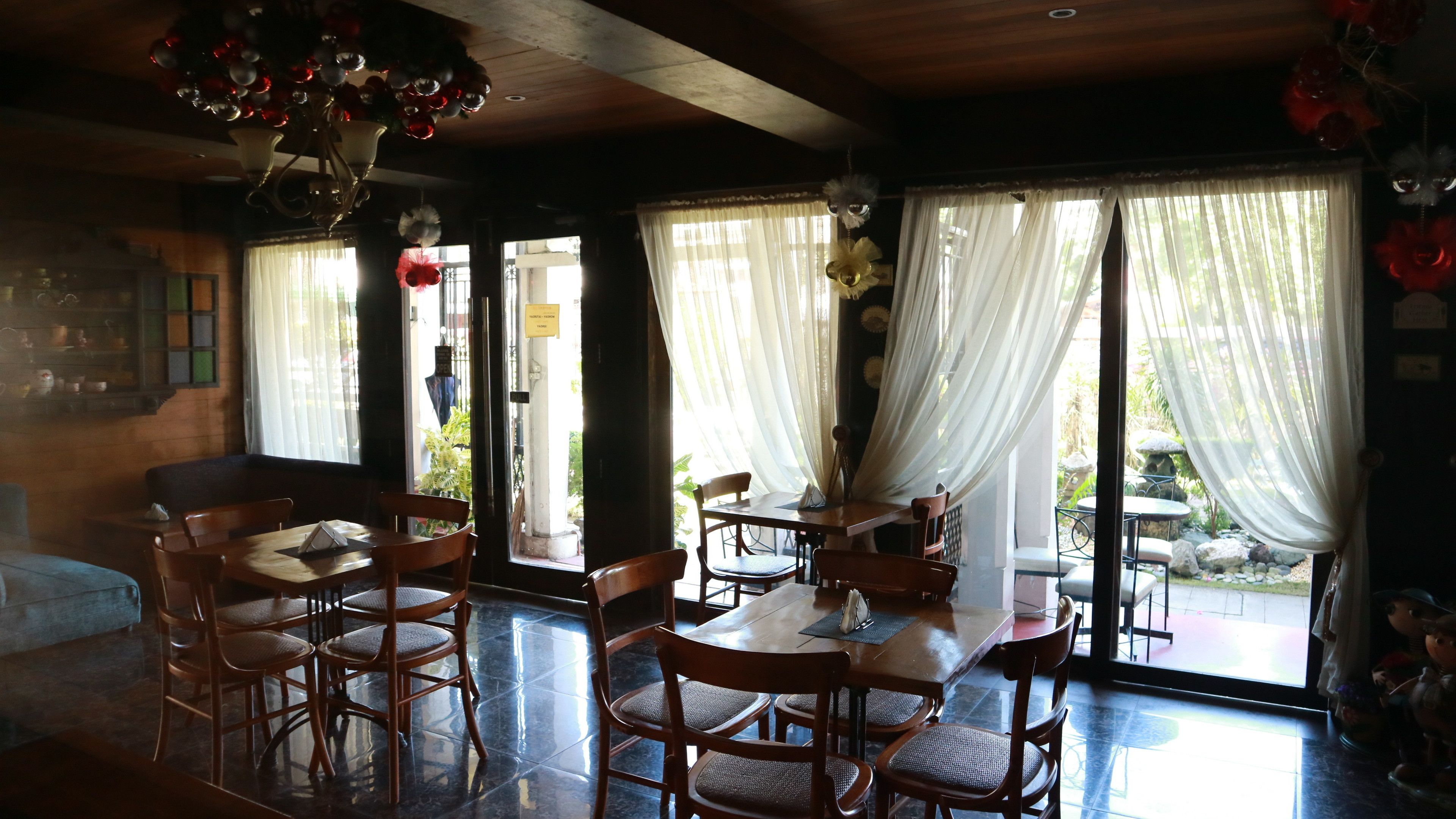 A dining area with wooden tables and chairs, large windows with sheer curtains, and a chandelier.
