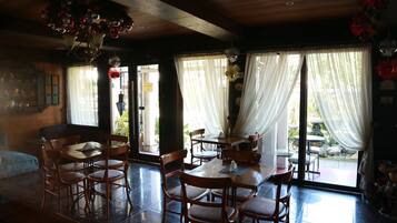 A dining area with wooden tables and chairs, large windows with sheer curtains, and a chandelier.