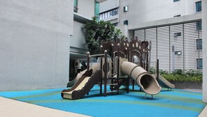 Un terrain de jeux avec des toboggans et des structures d’escalade, entouré d’une zone recouverte d’une moquette bleue, et un bâtiment à l’architecture moderne en arrière-plan.