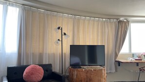 Uma sala de estar moderna com televisão de tela plana, mesa de centro de madeira, sofá preto com almofada rosa e um abajur.