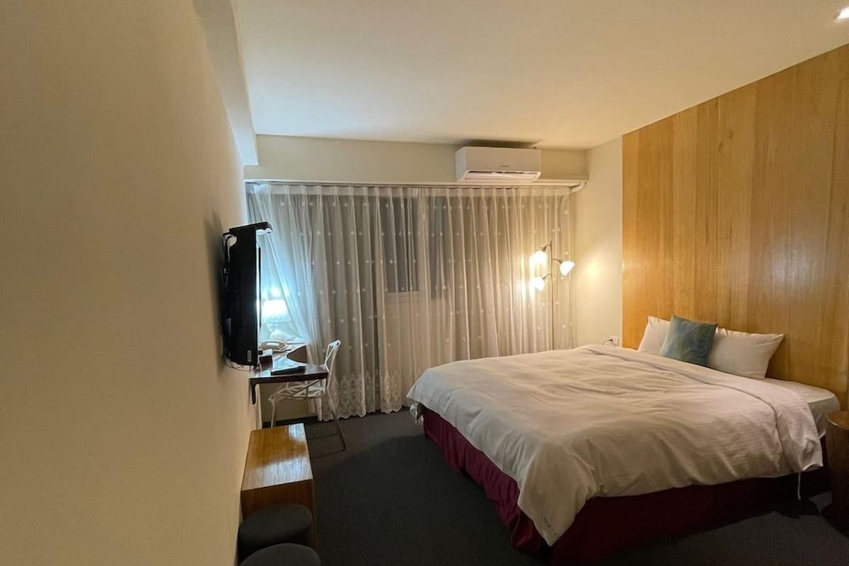 A hotel room with a bed, a desk with a chair, a television, and a window with curtains.