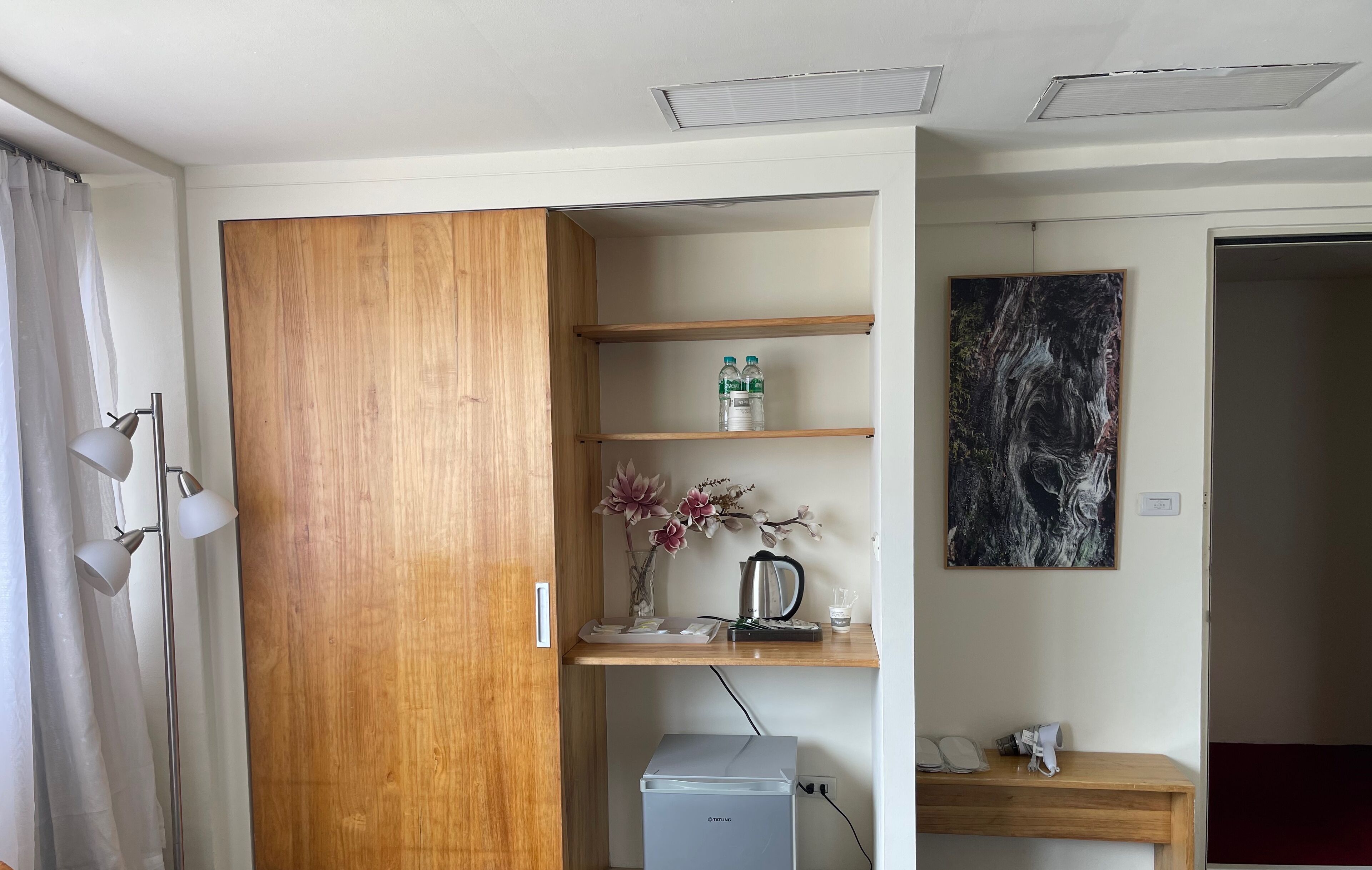 Una habitación con un armario de madera, un estante pequeño con botellas de agua, un florero con flores, una cafetera y un cuadro en la pared.
