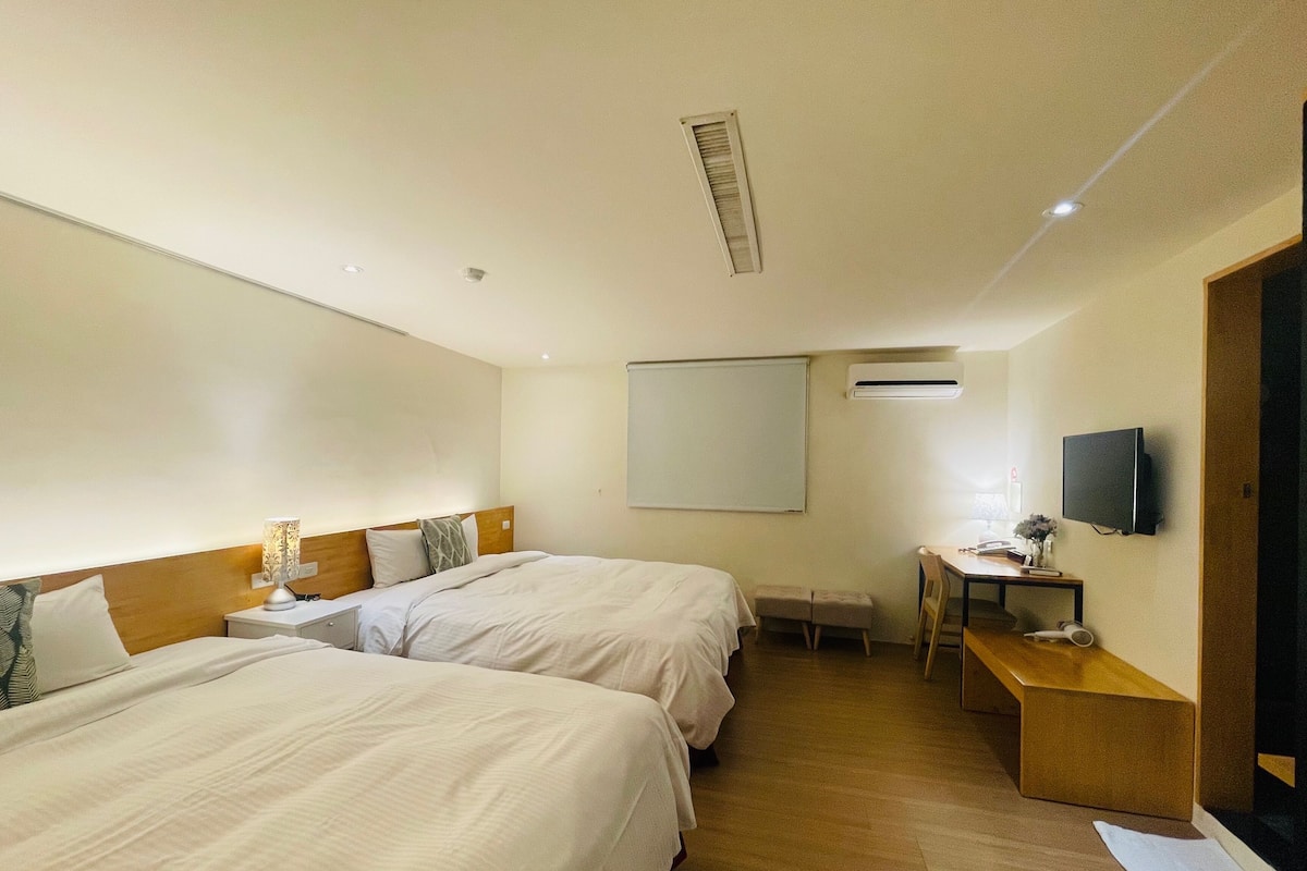 A hotel room with two beds, a desk with a chair, a TV, and an air conditioning unit.