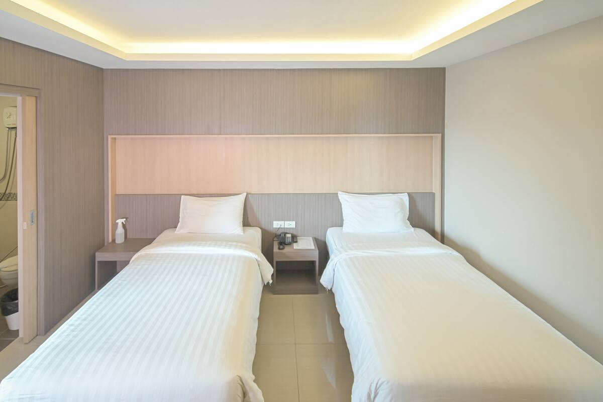 Habitación de hotel con dos camas individuales, cabecera de madera, mesita de noche con un teléfono y una botella, y suelo de baldosas.