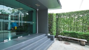 Un bâtiment moderne avec une façade en verre, un espace extérieur aménagé pour s’asseoir et une haie bien entretenue.