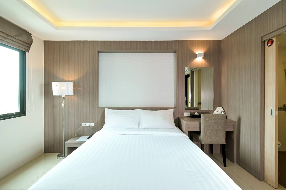 Habitación de hotel con una cama grande, una mesita de noche, un escritorio con silla, un espejo y una ventana con cortinas.