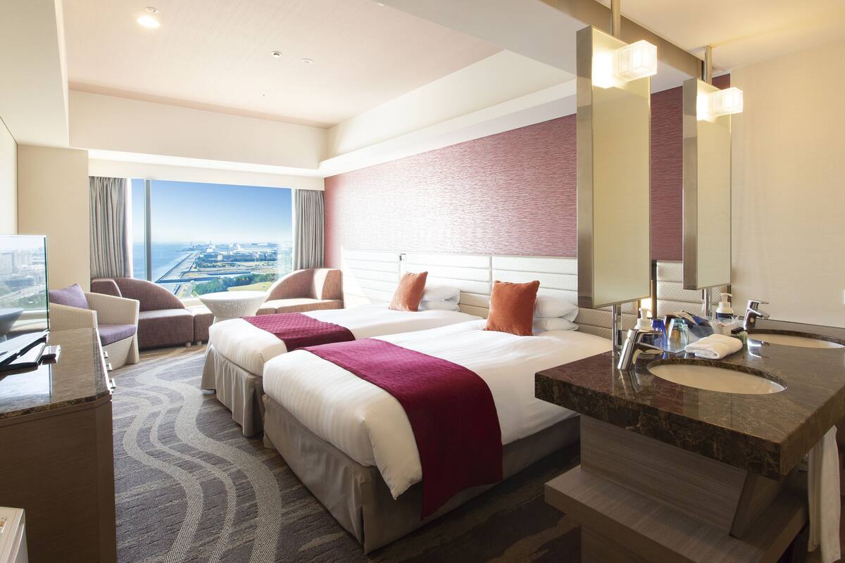 Habitación de hotel con dos camas, un ventanal grande con vistas a la ciudad, una zona de estar con sillas y un baño moderno con lavabo y espejo.