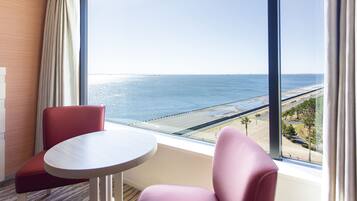A hotel room with a large window offering a view of the sea and a beach. There is a round table and two chairs.