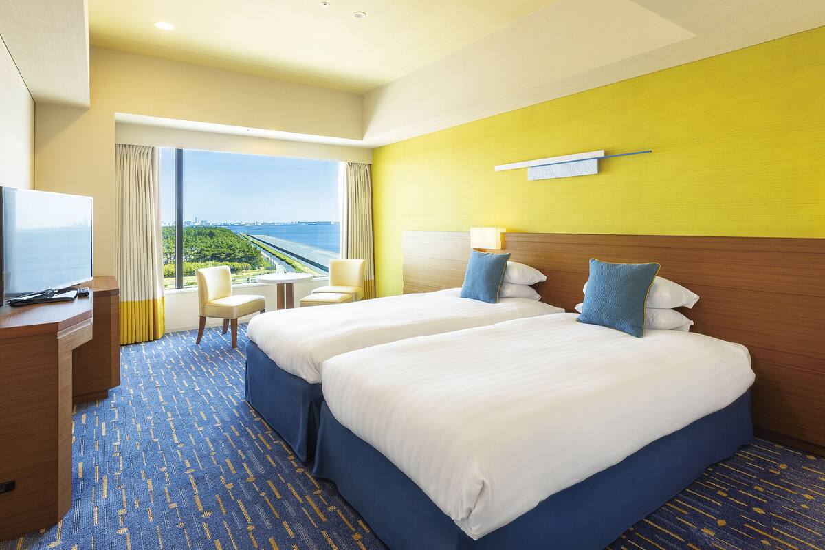Habitación de hotel con dos camas, televisión, escritorio y vistas a un puente y vegetación.