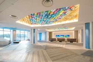 A spacious lobby with a modern design, featuring a unique ceiling installation, large windows, and a reception area with seating.