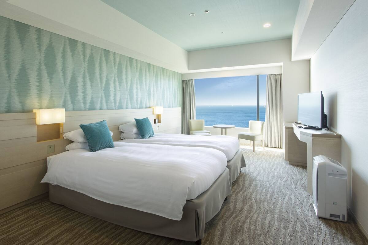 Habitación de hotel con dos camas, un ventanal con vistas al mar, un televisor de pantalla plana y una mesita.