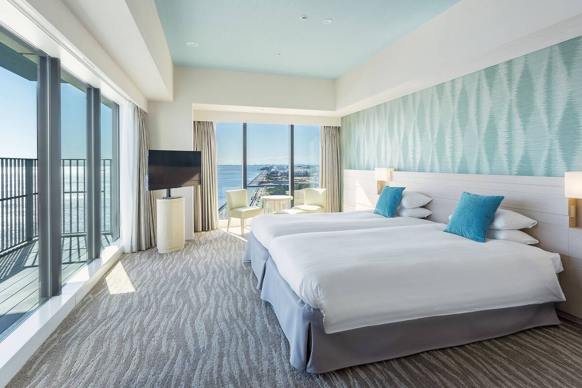 Habitación de hotel con una cama grande, televisión, balcón con vistas al agua y un pequeño rincón de descanso.