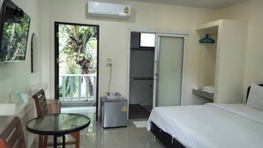 A hotel room with a bed, a small table, a chair, a balcony with a view of palm trees, and a sliding door leading to a shower area. - Baan  Kalaya Garden (Thep Krasattri)