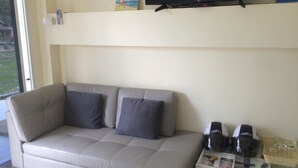 A modern living room with a beige sofa, a flat-screen TV mounted on the wall, and a small table with a coffee maker and cups. - Baan  Kalaya Garden (Thep Krasattri)